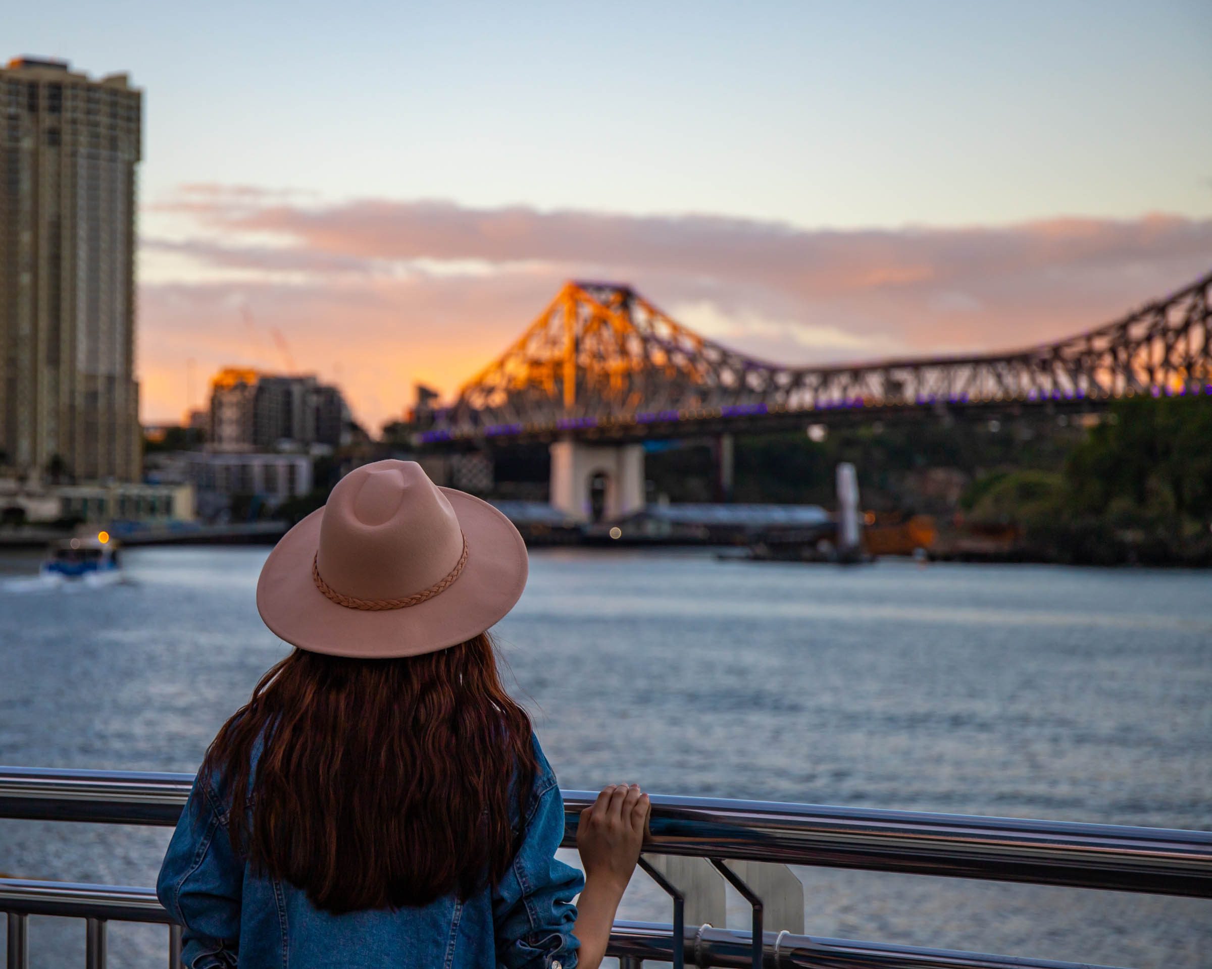 Individual wearing a hat looking at the Brisbane River and Story Bridge during sunset.